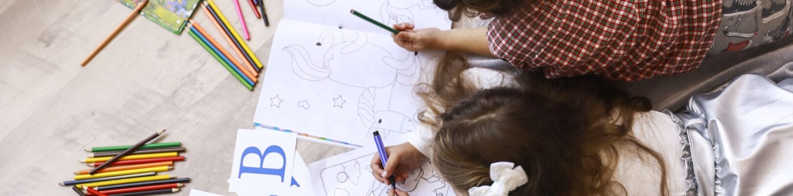 Top view of two tiny girls which are drawing in the coloring book laying on the floor on the blanket